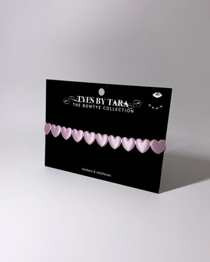 A pink heart-patterned bangle necklace displayed on a black branded card that reads 'TYES BY TARA THE BOWTYE COLLECTION'.