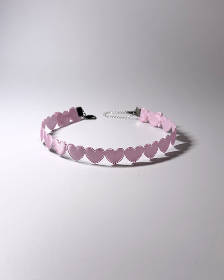 A light pink choker collar featuring a continuous row of small heart-shaped cutouts and a silver metal chain extender with a black clasp, shown on a neutral gray background.