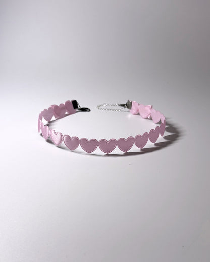 A light pink choker collar featuring a continuous row of small heart-shaped cutouts and a silver metal chain extender with a black clasp, shown on a neutral gray background.