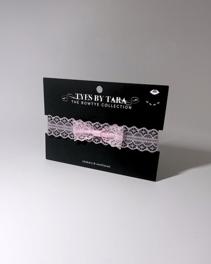 A light pink lace garter bow choker with a silver-tone chain extender with clasp, laid flat on a light gray background displayed on black TYES BY TARA branded black card