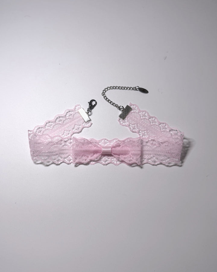 A light pink lace garter bow choker with a silver-tone chain extender with clasp, laid flat on a light gray background.