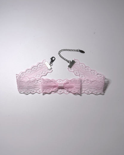 A light pink lace garter bow choker with a silver-tone chain extender with clasp, laid flat on a light gray background.