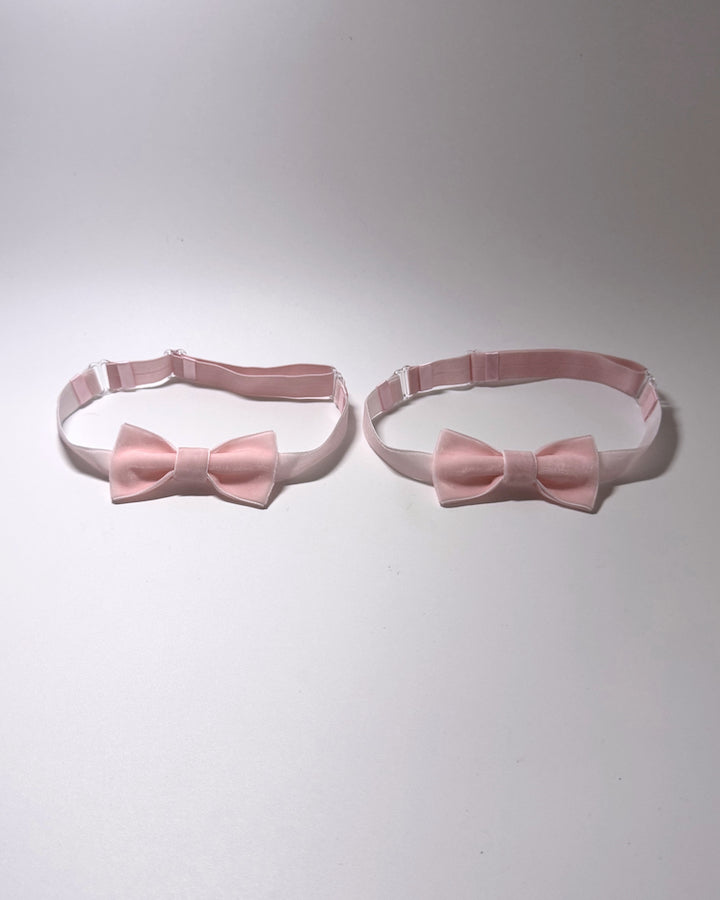 Two light pink satin bow leg garters with matching adjustable elastic straps, displayed side-by-side on a neutral light gray background.
