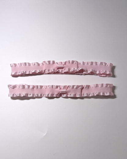 Two light pink ruffled satin garter straps with bow details, laid horizontally on a plain white background.