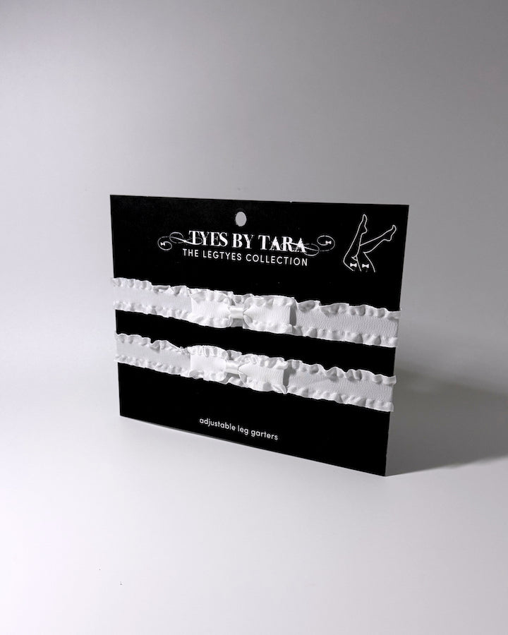 White Ruffled Bow Leg Garters on a black retail card displayed on light gray background with soft shadows