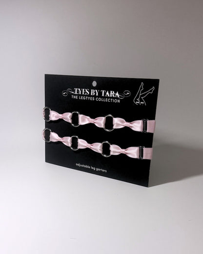 Two light pink satin bow leg garters with silver metal rings, displayed on a black card labeled 'TYES BY TARA THE LEGTYES COLLECTION'.