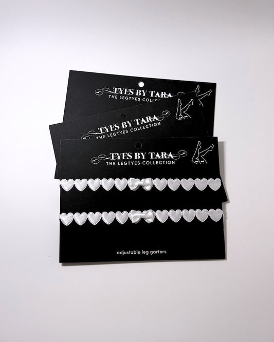 Three black display cards for 'Tyes By Tara The Legtyes Collection' holding two white heart-patterned adjustable leg garters, photographed on a plain light background.