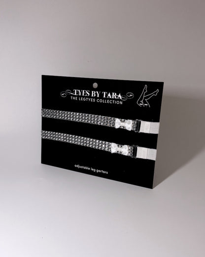 Sparkly silver white bow leg garters displayed on black retail card on a light gray background