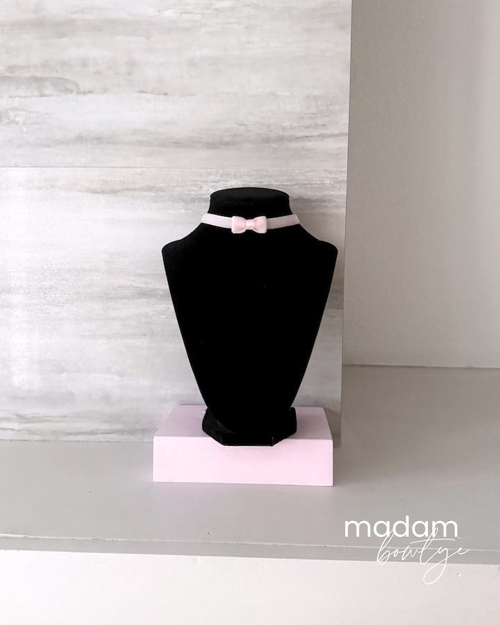 A light pink choker necklace with a small bow detail displayed on a black jewelry bust stand, set against a light gray textured wall.