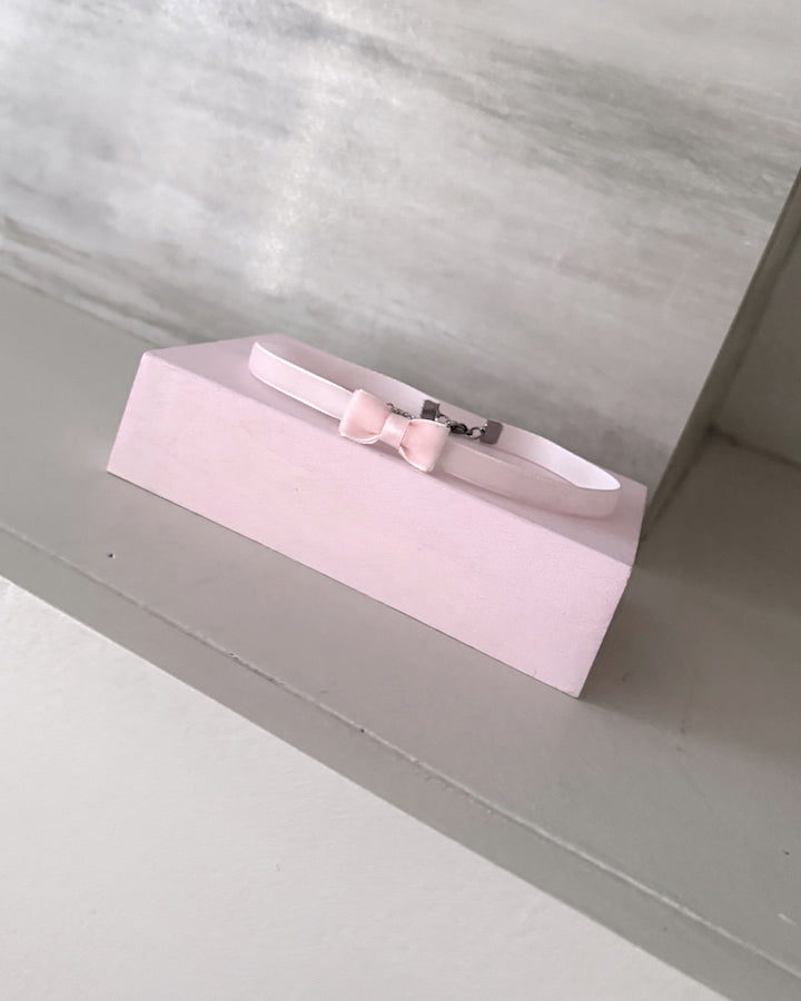 A light pink pet collar with a matching bow and silver buckle, displayed on a pink rectangular stand against a gray background.