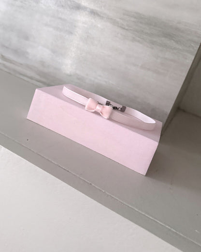 A light pink pet collar with a matching bow and silver buckle, displayed on a pink rectangular stand against a gray background.