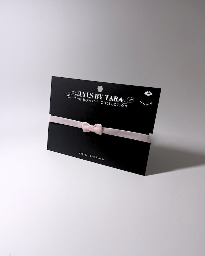 A pale pink velvet bow choker displayed on a black branded card from 'TYES by Tara - The Bowtye Collection'.