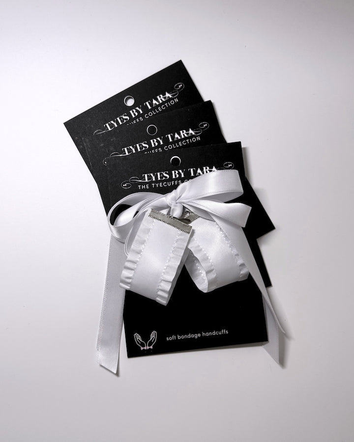 A white satin ruffled handcuffs with Tyes by Tara, presented on its original black packaging card with a white ribbon bow, shot on a light gray background.
