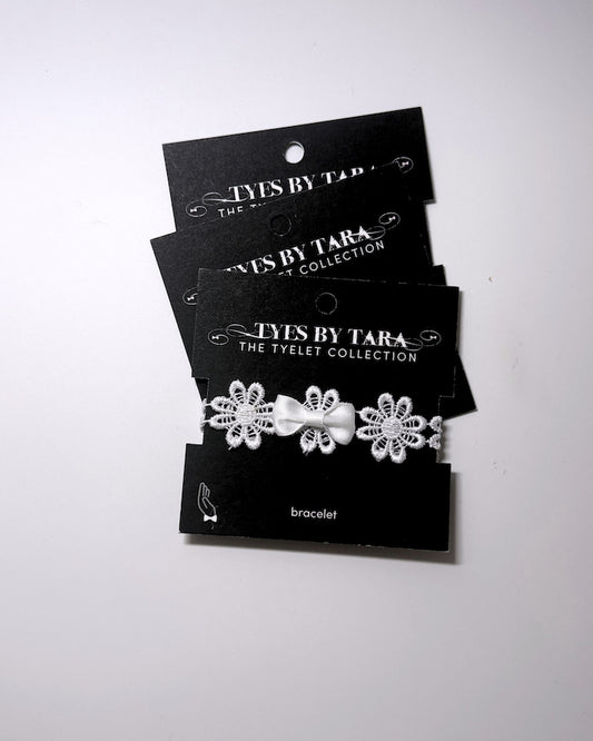 Three black jewelry display cards stacked on a white background, each holding a white flower bracelet with a small bow. The brand name 'TYES BY TARA' is printed on the cards.