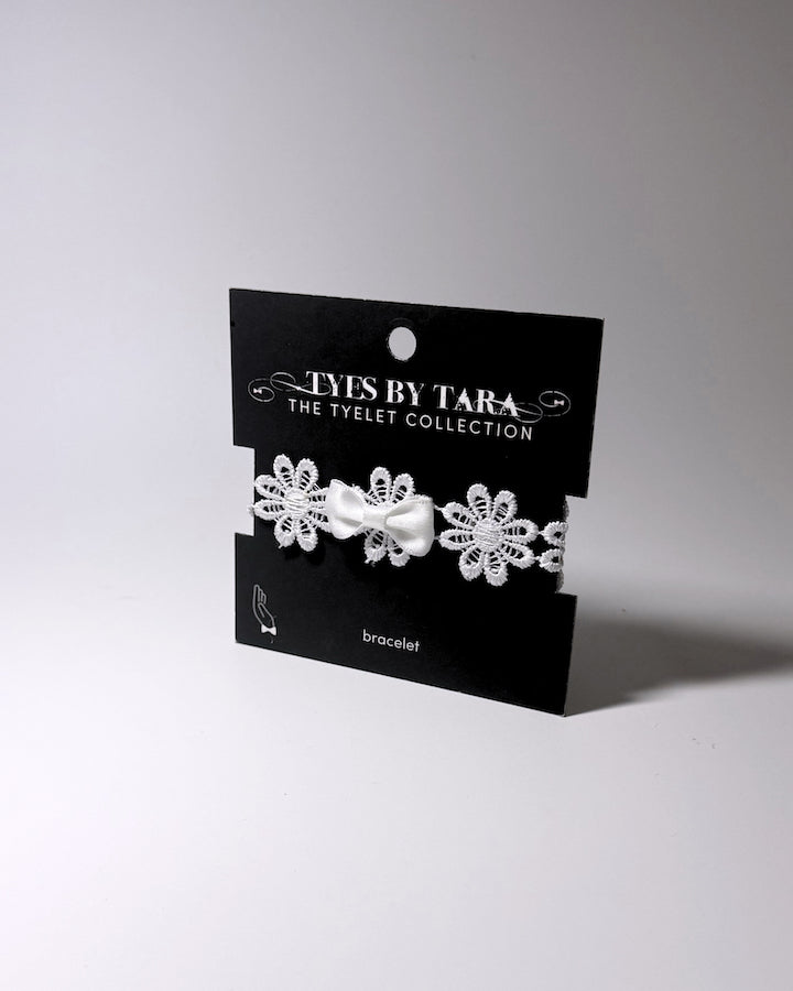 A white lace bracelet with a central satin bow, presented on a black display card labeled 'TYES BY TARA THE TYLET COLLECTION'.