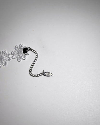 A close-up of a silver chain extender with a small oval tag, shown next to white floral lace trim on a plain light gray background.