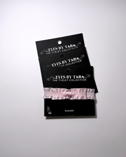 A pale pink ruffled bow bracelet displayed on a black retail card labeled 'TYES BY TARA THE TYELET COLLECTION' against a light gray studio background. 3 Pack shown