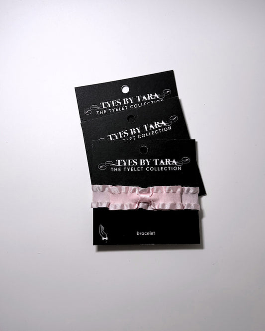 A pale pink ruffled bow bracelet displayed on a black retail card labeled 'TYES BY TARA THE TYELET COLLECTION' against a light gray studio background. 3 Pack shown