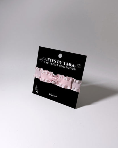 A pale pink ruffled bow bracelet displayed on a black retail card labeled 'TYES BY TARA THE TYELET COLLECTION' against a light gray studio background.