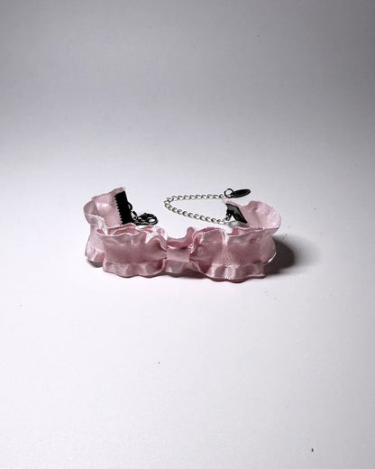 A light pink ruffled bow bracelet and a silver metal chain extender, photographed on a neutral gray background.