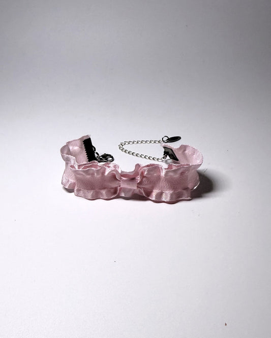 A light pink ruffled bow bracelet and a silver metal chain extender, photographed on a neutral gray background.