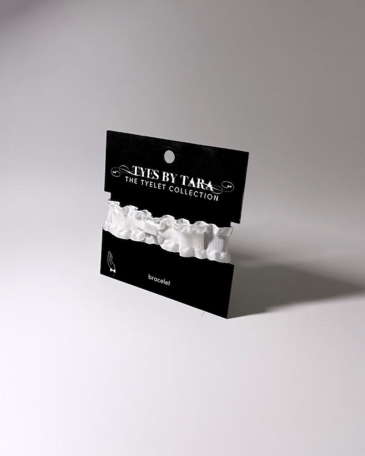 A white ruffled bow bracelet displayed on a black card labeled 'TYE BY TARA THE TYELET COLLECTION', photographed on a light gray studio background.