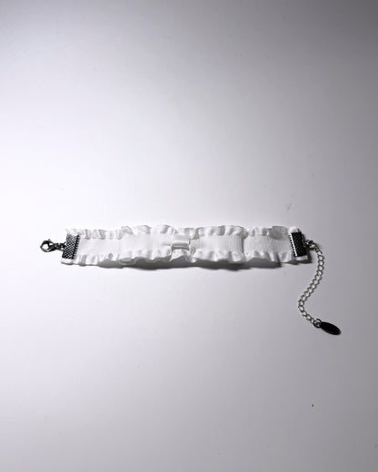 A white ruffled bow bracelet with stainless steel hardware, including a side clasp and an extender chain, laid flat on a light gray background.