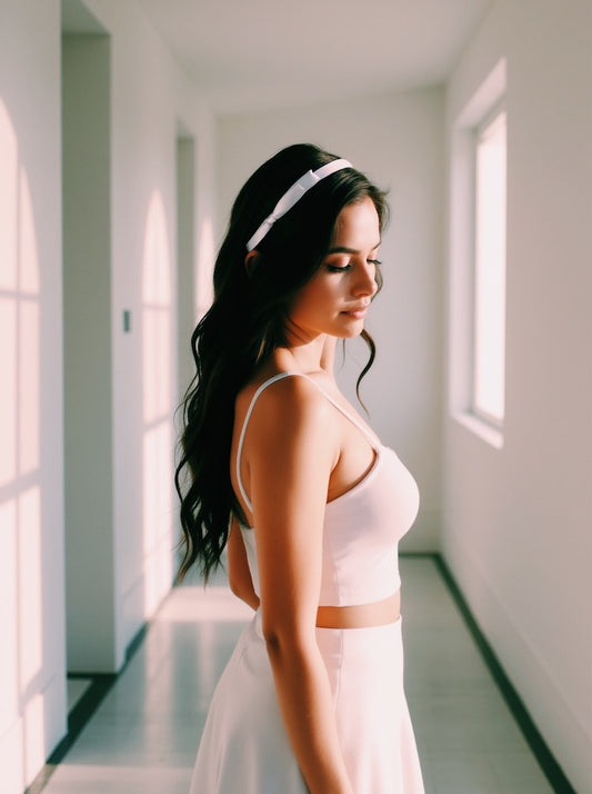 Woman wearing satin bow headband in a light pink top and white skirt standing in a sunlit room.
