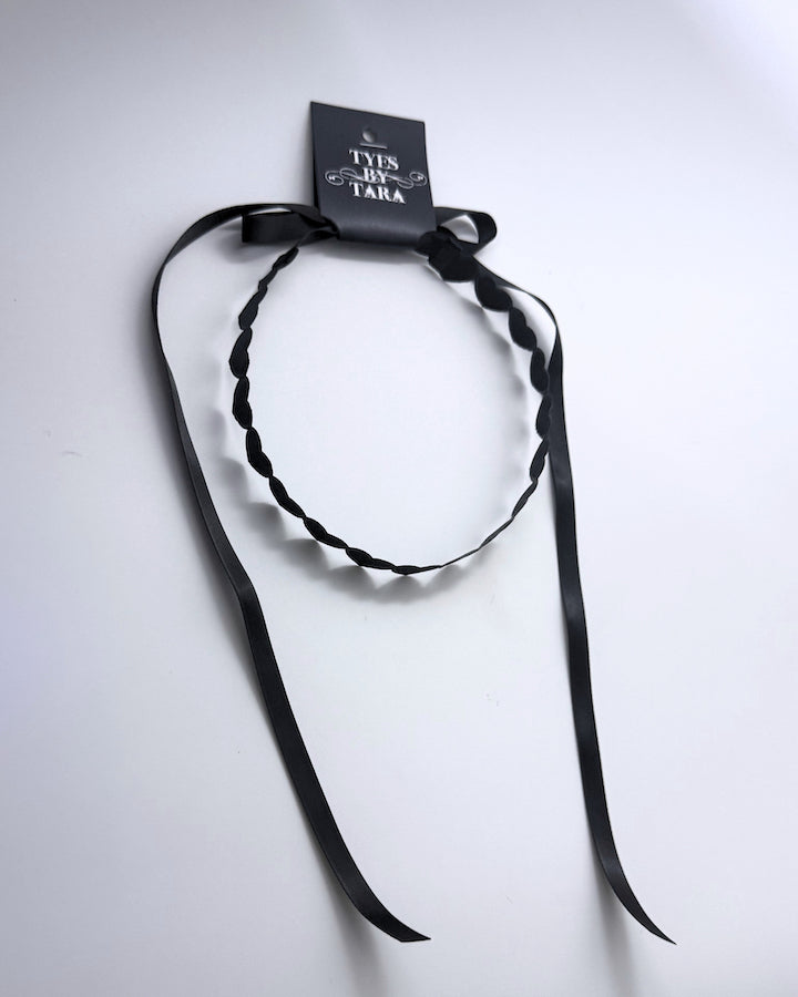 A black heart headband with long ribbon tails, attached to a black paper tag that reads 'TYES BY TARA', photographed on a plain white background.