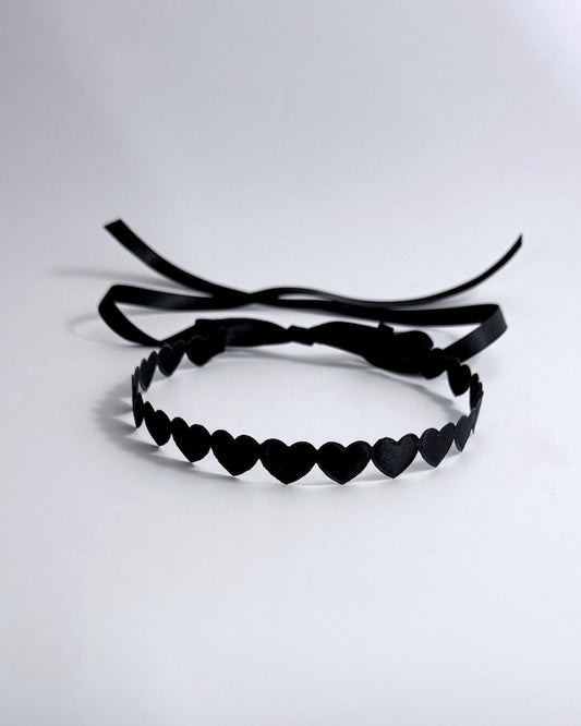 A black headband featuring a continuous row of small heart-shaped cutouts and long ribbon ties, displayed on a clean white background.
