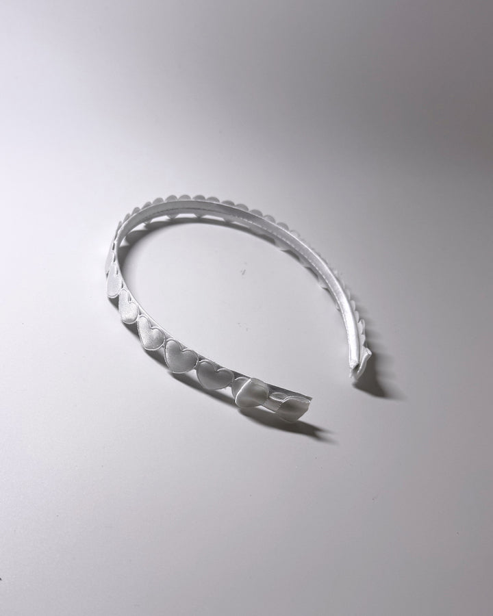 A white plastic headband with a scalloped hearts and a matching bow on ends of headband, photographed on a neutral light gray background.