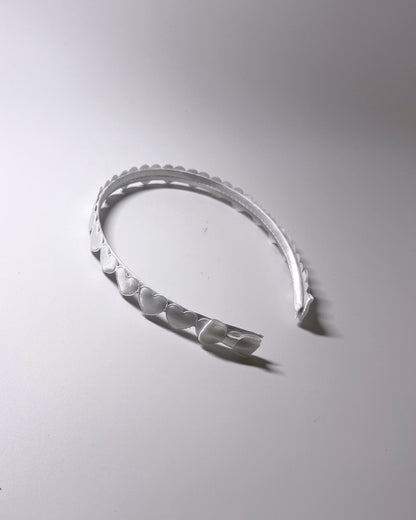 A white plastic headband with a scalloped hearts and a matching bow on ends of headband, photographed on a neutral light gray background.