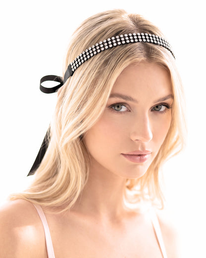 A blonde woman wearing a black headband embellished with sparkling rhinestones and a black ribbon bow, photographed against a bright white background.
