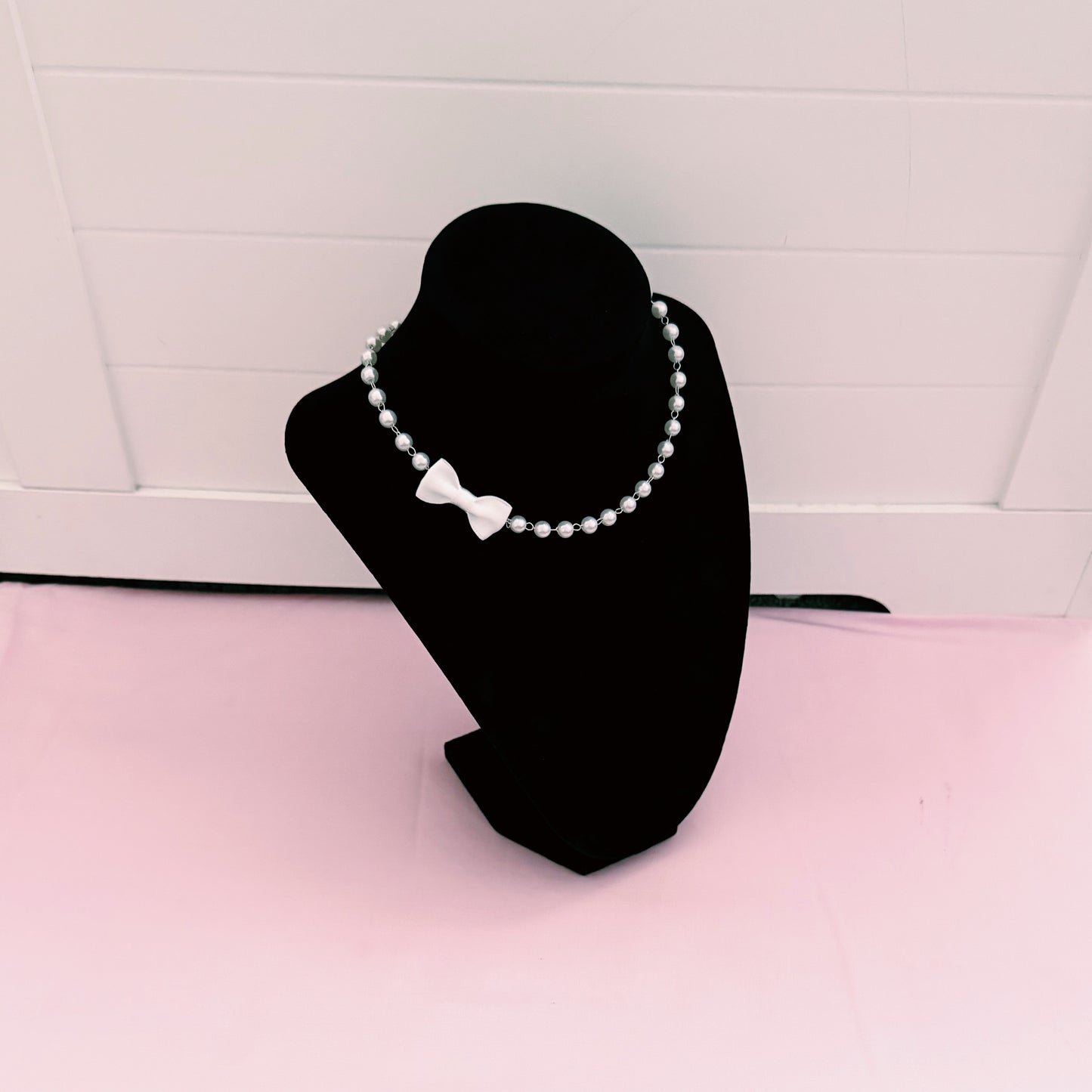 Pearl white bow necklace on a black mannequin bust against a white tiled wall.