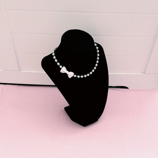 Pearl white bow necklace on a black mannequin bust against a white tiled wall.