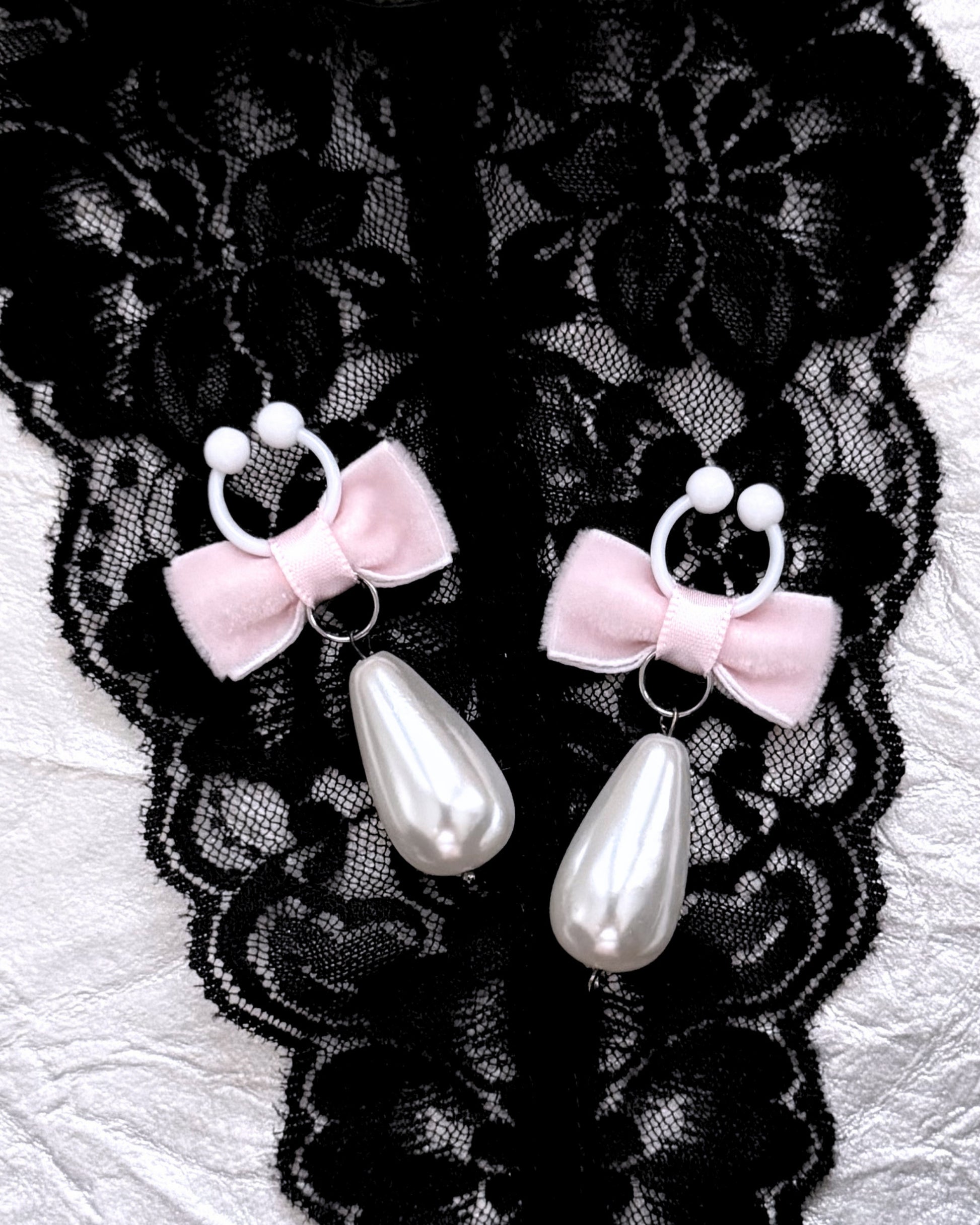 A pair of nipple clamps with pale pink velvet bows, white flexible clamps with teardrop-shaped white faux pearls, displayed on black lace fabric over a white textured surface.