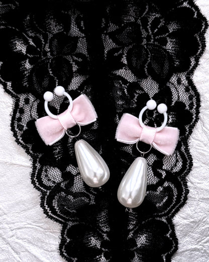 A pair of nipple clamps with pale pink velvet bows, white flexible clamps with teardrop-shaped white faux pearls, displayed on black lace fabric over a white textured surface.