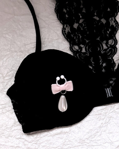 A pair of nipple clamps with pale pink velvet bows, white flexible clamps with teardrop-shaped white faux pearls, displayed on black lace fabric over a black lacy bra