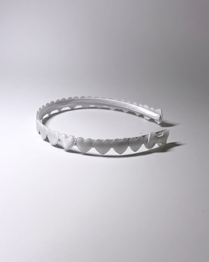 A white plastic headband with a scalloped hearts and a matching bow on ends of headband, photographed on a neutral light gray background side view
