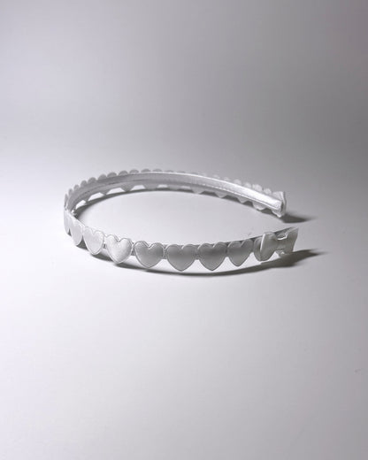 A white plastic headband with a scalloped hearts and a matching bow on ends of headband, photographed on a neutral light gray background side view