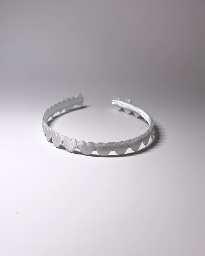 A white plastic headband with a scalloped hearts and a matching bow on ends of headband, photographed on a neutral light gray background showing the top of the hairband