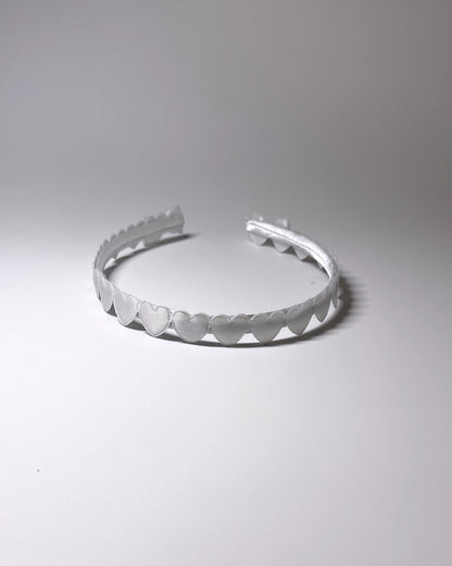 A white plastic headband with a scalloped hearts and a matching bow on ends of headband, photographed on a neutral light gray background showing the top of the hairband